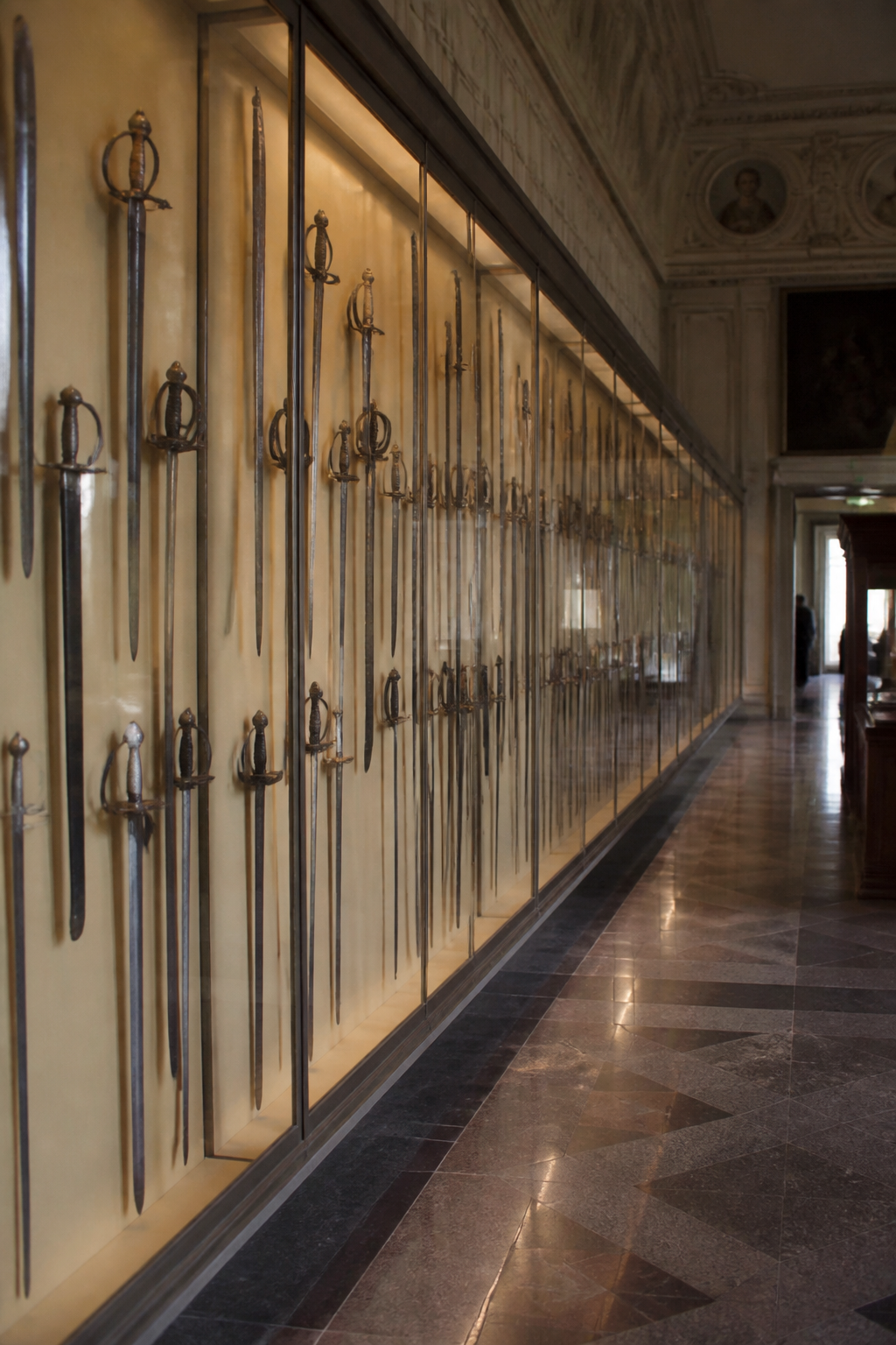 Long museum gallery lined with glass cases of edged weapons, swords mounted vertically in rows under even lighting, with a polished stone floor running the length of the room